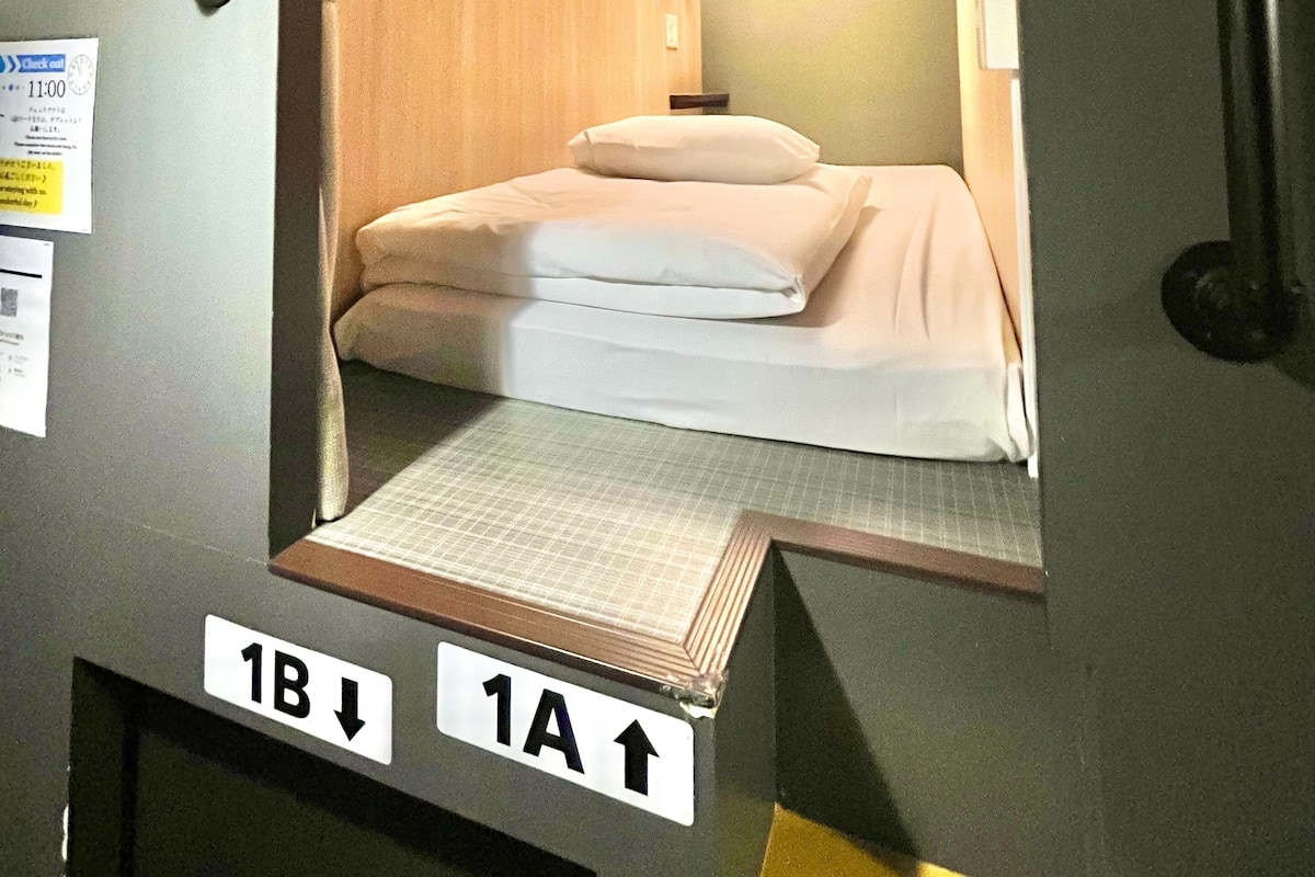 Una habitación pequeña con cama individual, cabecera de madera, lámpara de noche y un ventanal. Se observan señales direccionales que indican las habitaciones 1A y 1B.