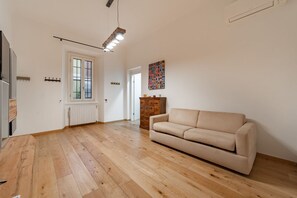 Ein geräumiger Raum mit Holzboden, einem beigefarbenen Sofa, einem Heizkörper und einer Tür zu einem anderen Zimmer.