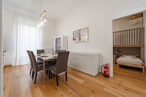 Ein modernes, helles Zimmer mit Esstisch, Stühlen, einem weißen Schrank, einem Bett und einem Feuerlöscher.