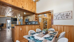 Un intérieur en bois comprenant un coin repas avec une table agrémentée de sets de table sur le thème de la forêt, un coin cuisine doté d’appareils modernes et une œuvre d’art accrochée au mur.