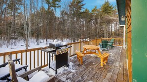 Une terrasse en bois avec un barbecue, des sièges d’extérieur et une vue sur une forêt enneigée.
