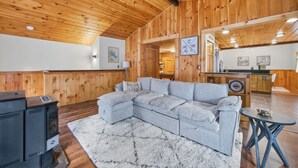 Un intérieur de chalet chaleureux avec un plafond en bois, un canapé d’angle, une cheminée et un coin cuisine.