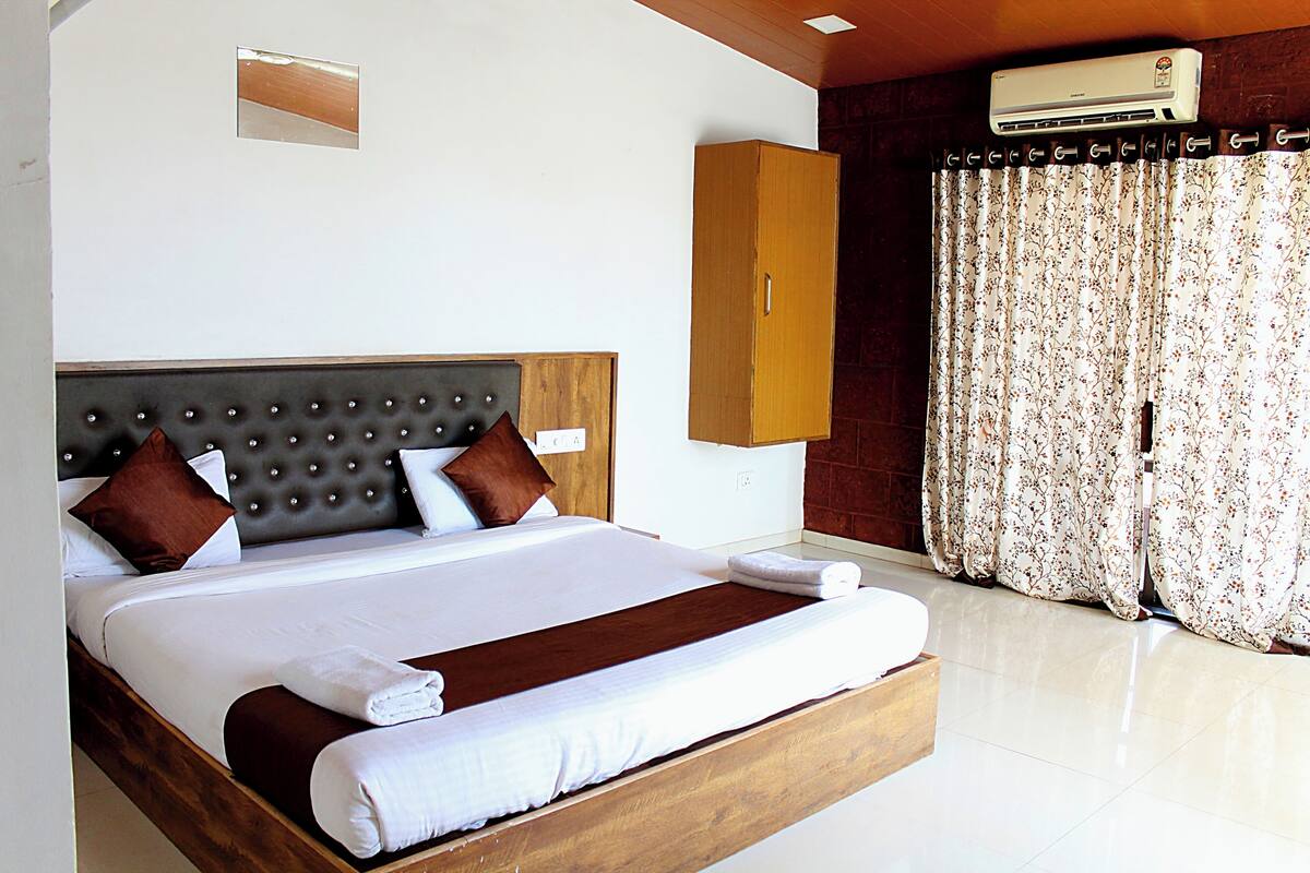 Quarto de hotel com uma cama grande, cabeceira de madeira, guarda-roupa e uma janela com cortinas florais.