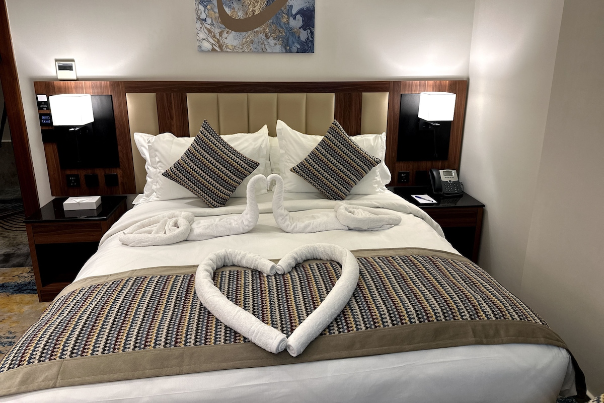 A hotel room with a neatly made bed, two pillows, a heart-shaped towel arrangement, and a wall-mounted artwork.