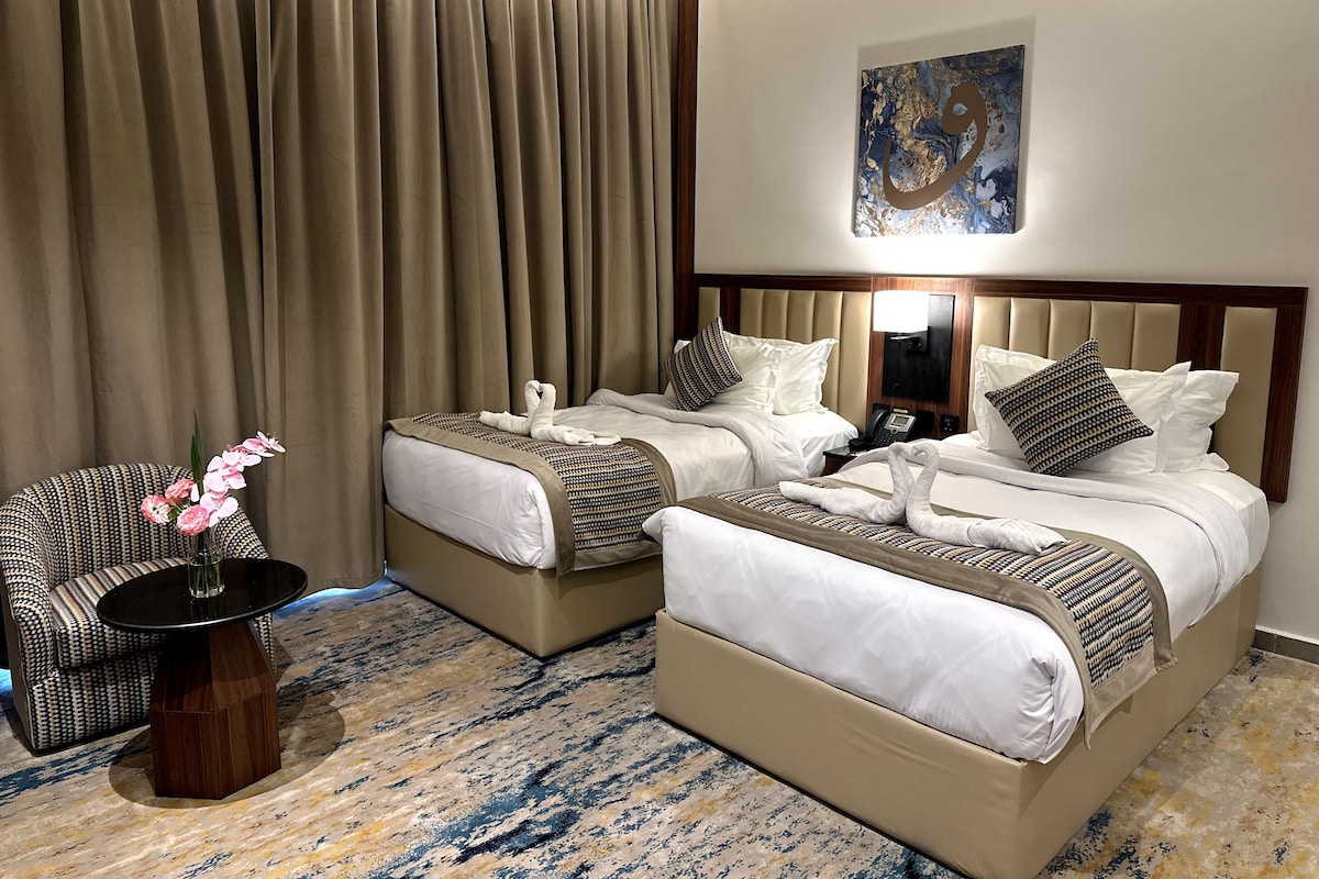 A hotel room with two beds, a chair, a small table, and a painting on the wall.