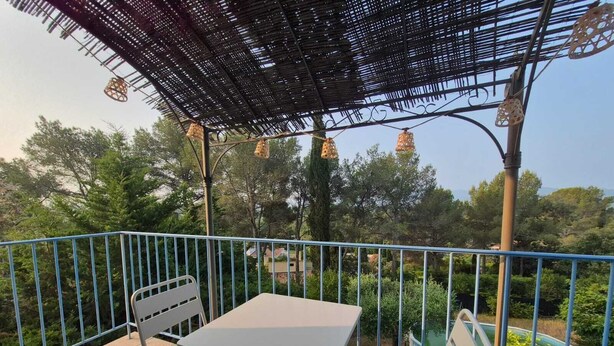 Un balcón con mesa y sillas, rodeado de una barandilla azul, con vistas a un jardín con árboles y una piscina.