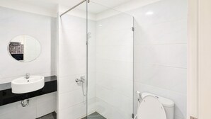 A modern bathroom with a glass shower enclosure, a round mirror, a white sink, and a toilet. - Altuz Apartment Seturan – Private City Studio with Sky Pool Access (Sleman)