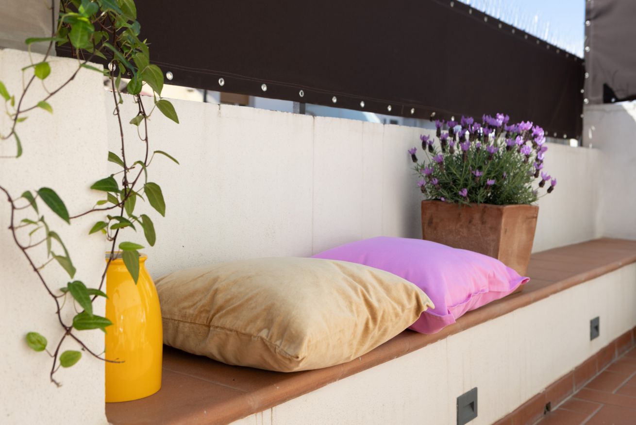 Un balcón con un cojín color beige, un jarrón amarillo y una planta en maceta con flores moradas.