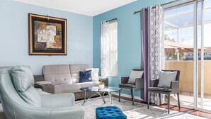 Una sala moderna con sofá, sillón y reposapiés, una mesa de centro de vidrio, y vistas a través de una puerta corrediza de cristal.
