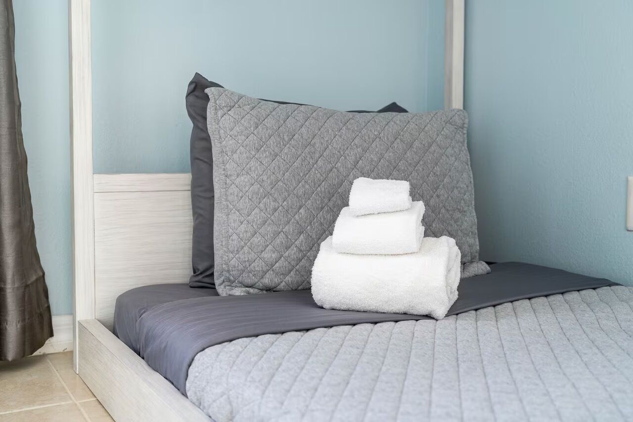 A neatly made bed with grey bedding and pillows, a stack of white towels, and a light blue wall in the background.