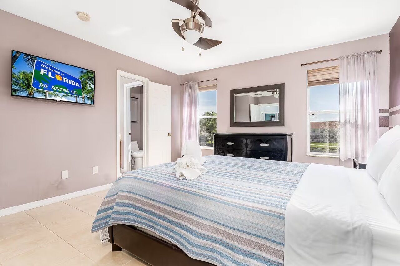 Una camera da letto con un letto, una ventola a soffitto, un televisore che mostra un cartello di benvenuto in Florida, un comò e una finestra con tende.