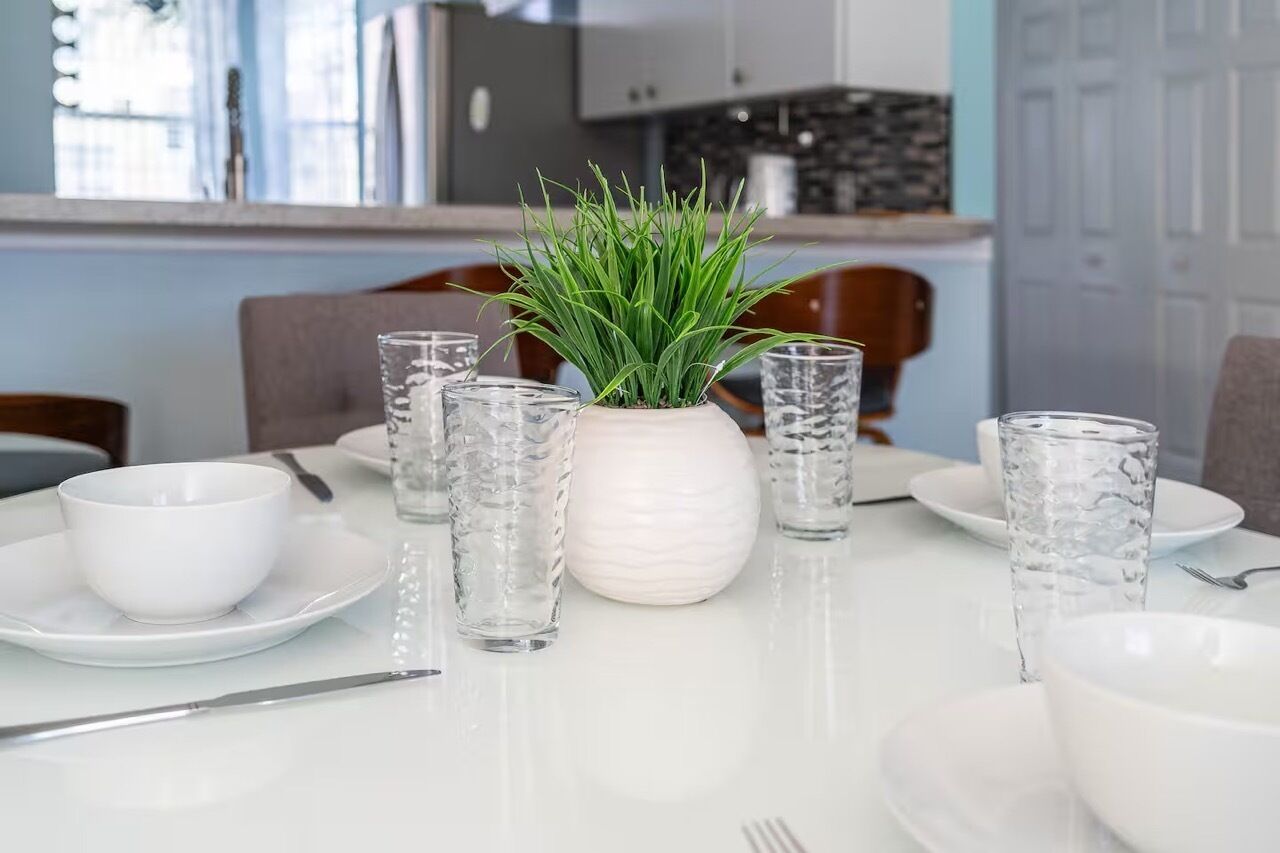 A dining table set with white plates, bowls, and clear glasses, with a potted plant in the center.