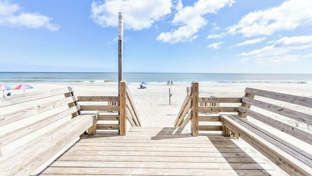 Ein hölzerner Steg führt zu einem Strand, mit einem Holzpfosten, auf dem "11" steht, und einem sandigen Strand im Hintergrund.