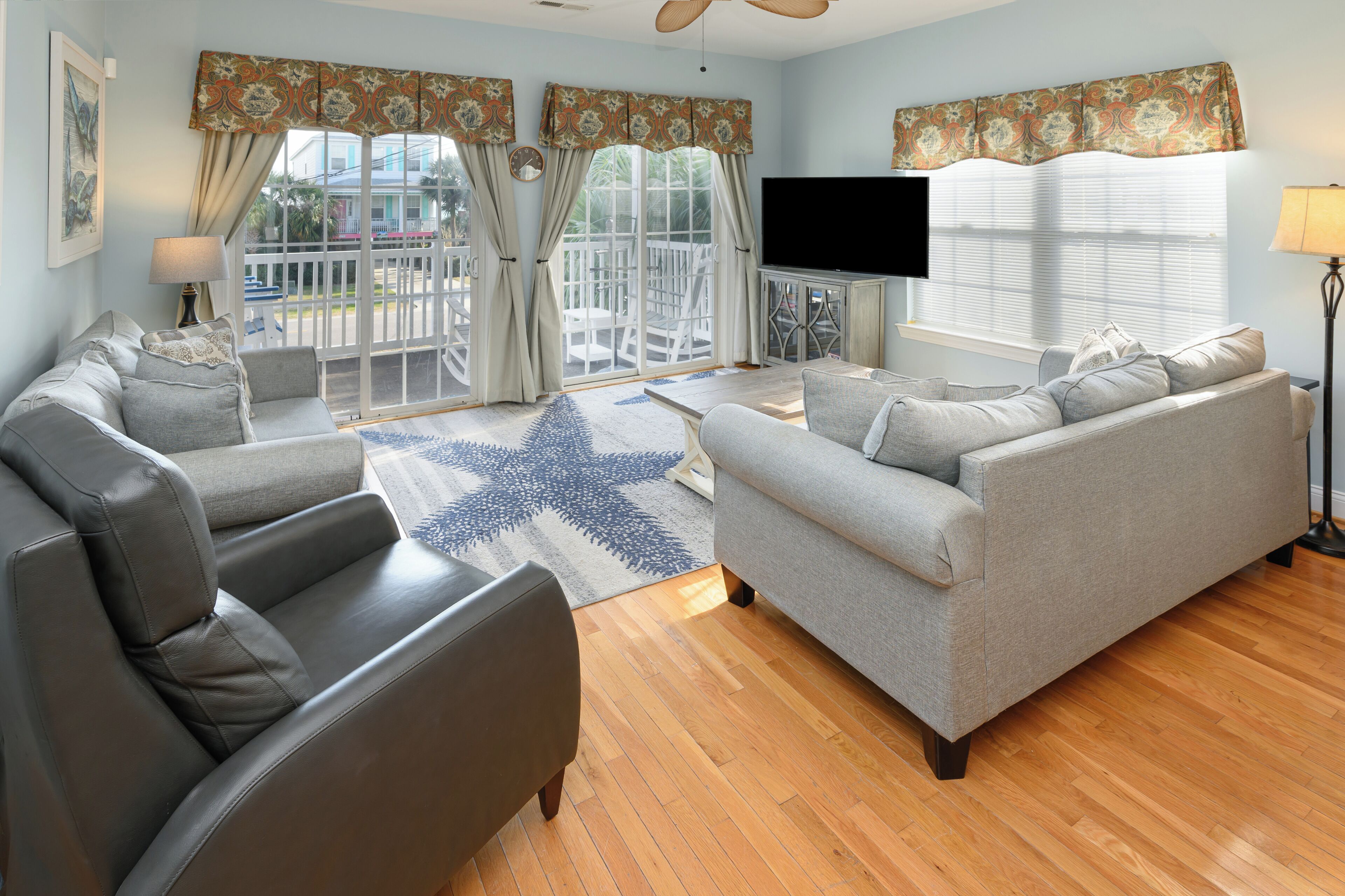 A living room with a grey sofa, a coffee table, and a TV. There are curtains, a lamp, and a view of the outside.