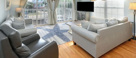 A living room with a grey sofa, a coffee table, and a TV. There are curtains, a lamp, and a view of the outside.