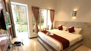 A hotel room with a large bed, a TV, a desk with a computer and a water bottle, and a view of the outdoors. - Ozone Hill Resorts (Yercaud)