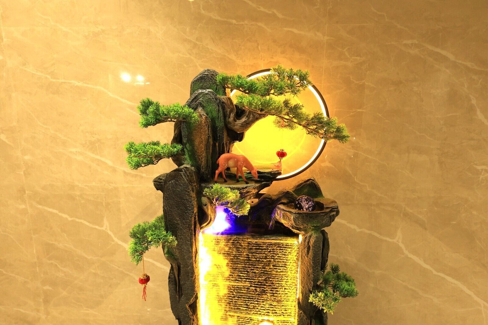 A decorative indoor fountain with a deer figurine, surrounded by greenery and illuminated by a circular light.