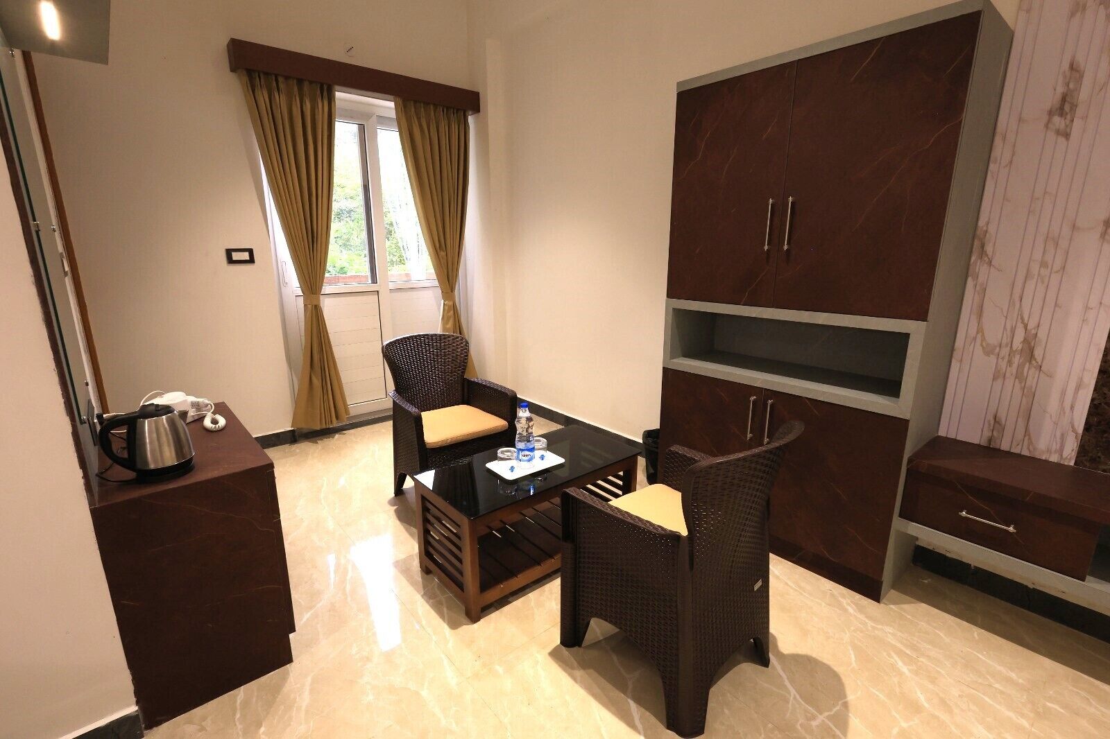 A compact room with a dining table, two chairs, a kettle, a water bottle, and a cabinet with drawers.