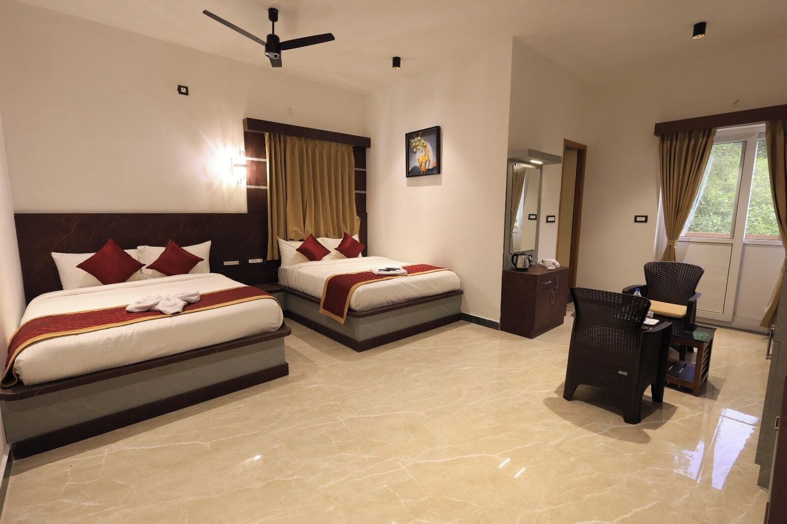 A hotel room with two beds, a sitting area with a chair and table, a ceiling fan, and a window with curtains.