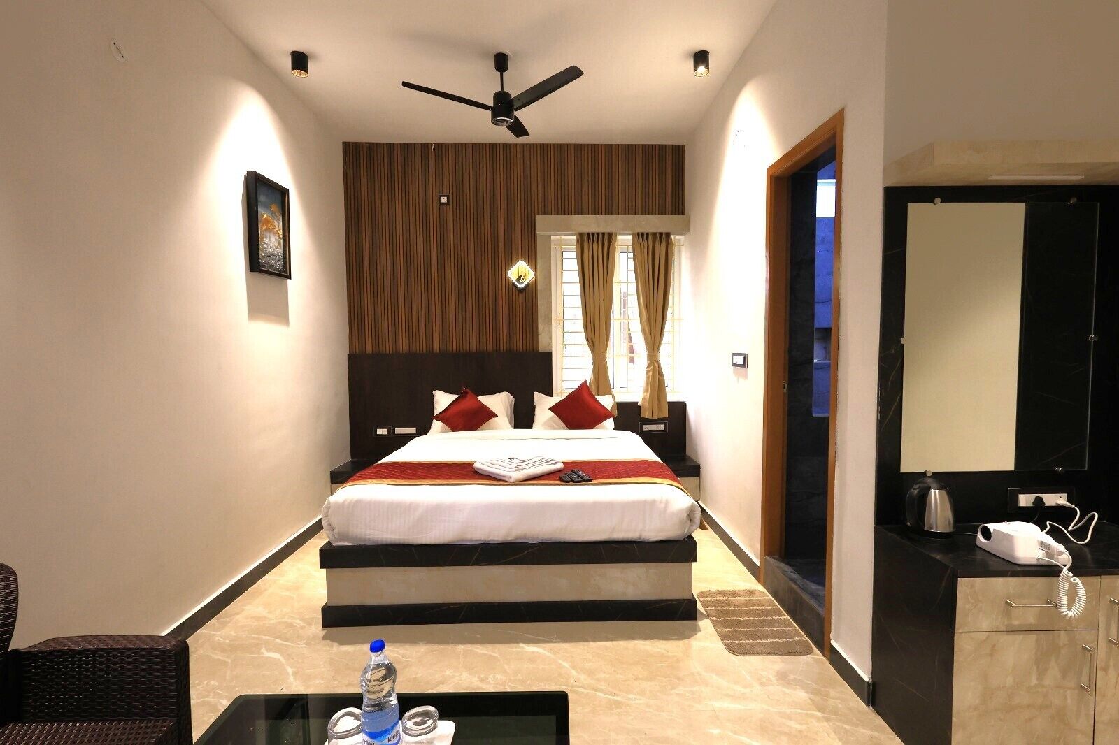 A hotel room with a bed, a desk with a kettle, a mirror, and a ceiling fan.