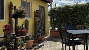 Ein gelbes Gebäude mit einer Terrasse, die einen Tisch und Stühle bietet, umgeben von Topfpflanzen und einem Kaktus.