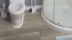 Een badkamer met toilet, een douchecabine van glas, een plank met handdoeken en een toiletrolhouder.
