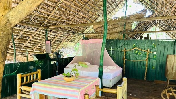 A thatched-roof outdoor room with a bed, a table with a striped tablecloth, and two chairs.