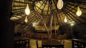 A rustic dining area with wooden tables and chairs, a thatched roof, and hanging light fixtures.