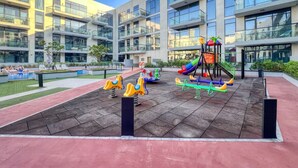 A playground with swings, slides, and climbing structures, surrounded by a modern building with large windows and balconies. - Spacious 1BR Sleeps 4+Pool, Gym & Rooftop Terrace (دبي)
