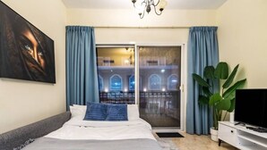 A bedroom with a bed, a TV, a balcony with a view of buildings, and curtains. - Spacious 1-BR Opposite Mall with Huge Pool & Gym (Dubai)