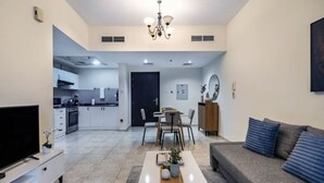 A modern living room with a flat-screen TV, a coffee table with decorative items, a grey sofa with blue cushions, and a dining area with chairs and a table. - Spacious 1-BR Opposite Mall with Huge Pool & Gym (Dubai)