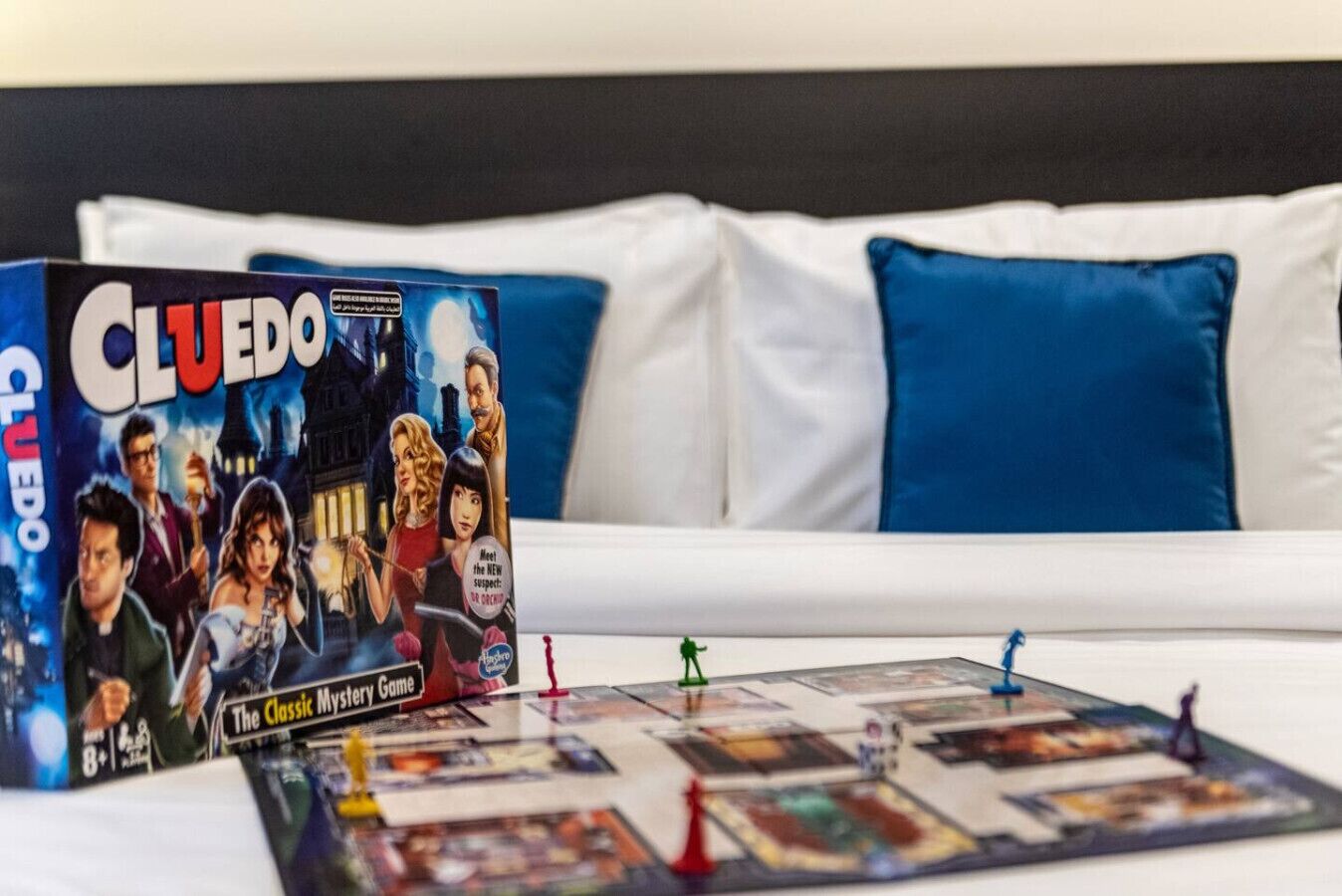 A Cluedo board game set on a bed with a white and blue pillow.