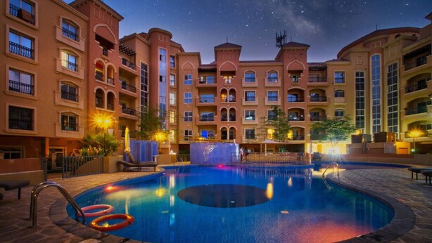 A swimming pool surrounded by multi-story buildings with balconies, under a night sky with stars. - Spacious 1-BR Opposite Mall with Huge Pool & Gym (Dubai)