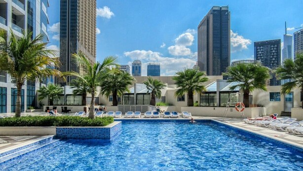 Ein Swimmingpool mit Liegestühlen, Palmen und modernen Gebäuden im Hintergrund.