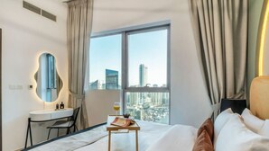 Ein Hotelzimmer mit einem Bett, einem Schreibtisch, einem Sessel und einem Spiegel, mit Blick auf die Stadt durch große Fenster.