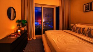 A hotel room with a bed, a nightstand, a potted plant, and a view of a cityscape at night. - Family & Group Friendly,  Near Beach Metro & Marina (Dubai)