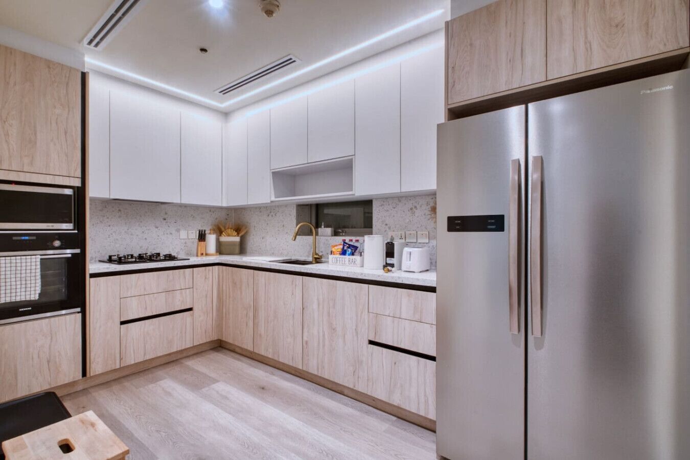 Een moderne keuken met lichte houten kastjes, een roestvrijstalen koelkast, een inbouwoven en een koffiehoek.