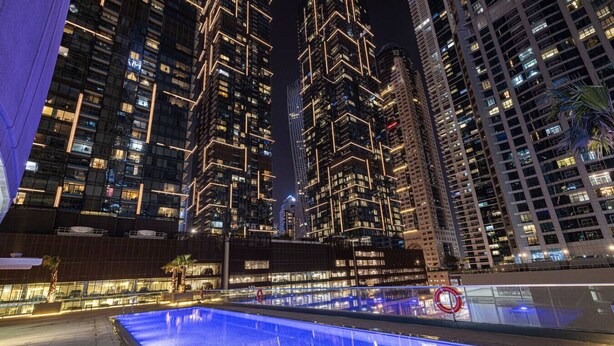 A nighttime view of a modern high-rise building with illuminated windows and a pool area with a blue-lit water feature. - Family & Group Friendly,  Near Beach Metro & Marina (Dubai)