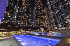 A nighttime view of a modern high-rise building with illuminated windows and a pool area with a blue-lit water feature. - Family & Group Friendly,  Near Beach Metro & Marina (Dubai)