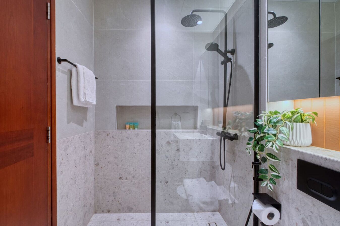Un baño moderno con cabina de ducha de vidrio, paredes de mármol, puerta de madera, un perchero, estante con artículos de aseo y una planta en maceta.
