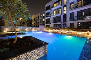 A swimming pool with lounge chairs and a tree, surrounded by modern buildings with balconies. - Urban Escape 1BR w/ pool+rooftop (دبي)