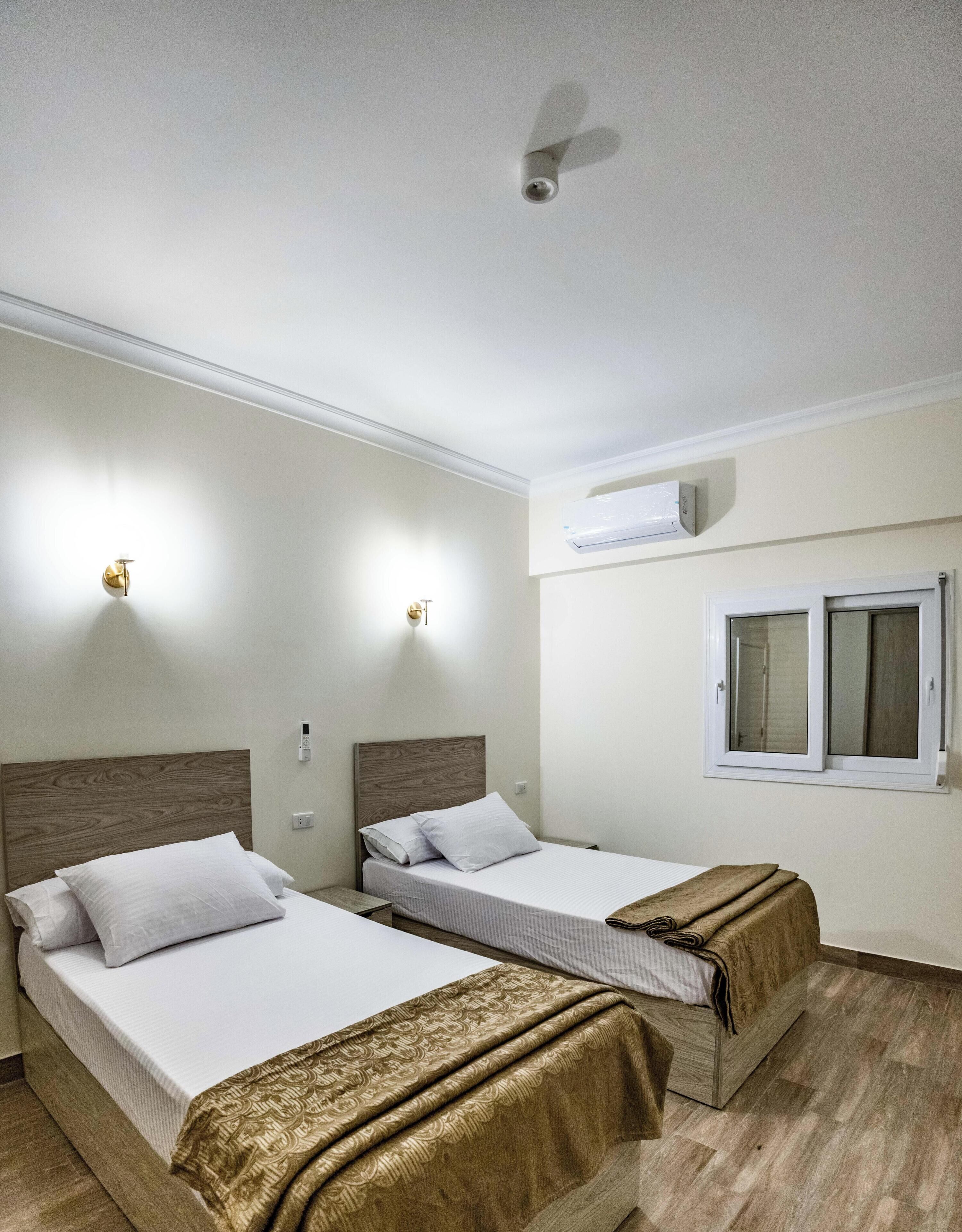 A hotel room with two single beds, a ceiling fan, an air conditioning unit, and a window with blinds.