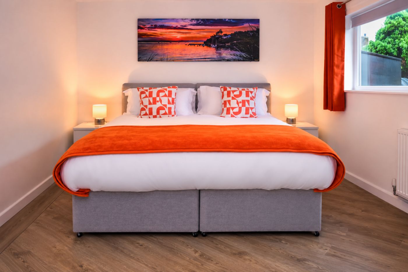 Ein Bett mit grauem Gestell, weißer Bettwäsche und orangefarbenen Akzenten, flankiert von zwei Nachttischlampen, in einem Zimmer mit Holzboden und einem Gemälde über dem Bett.