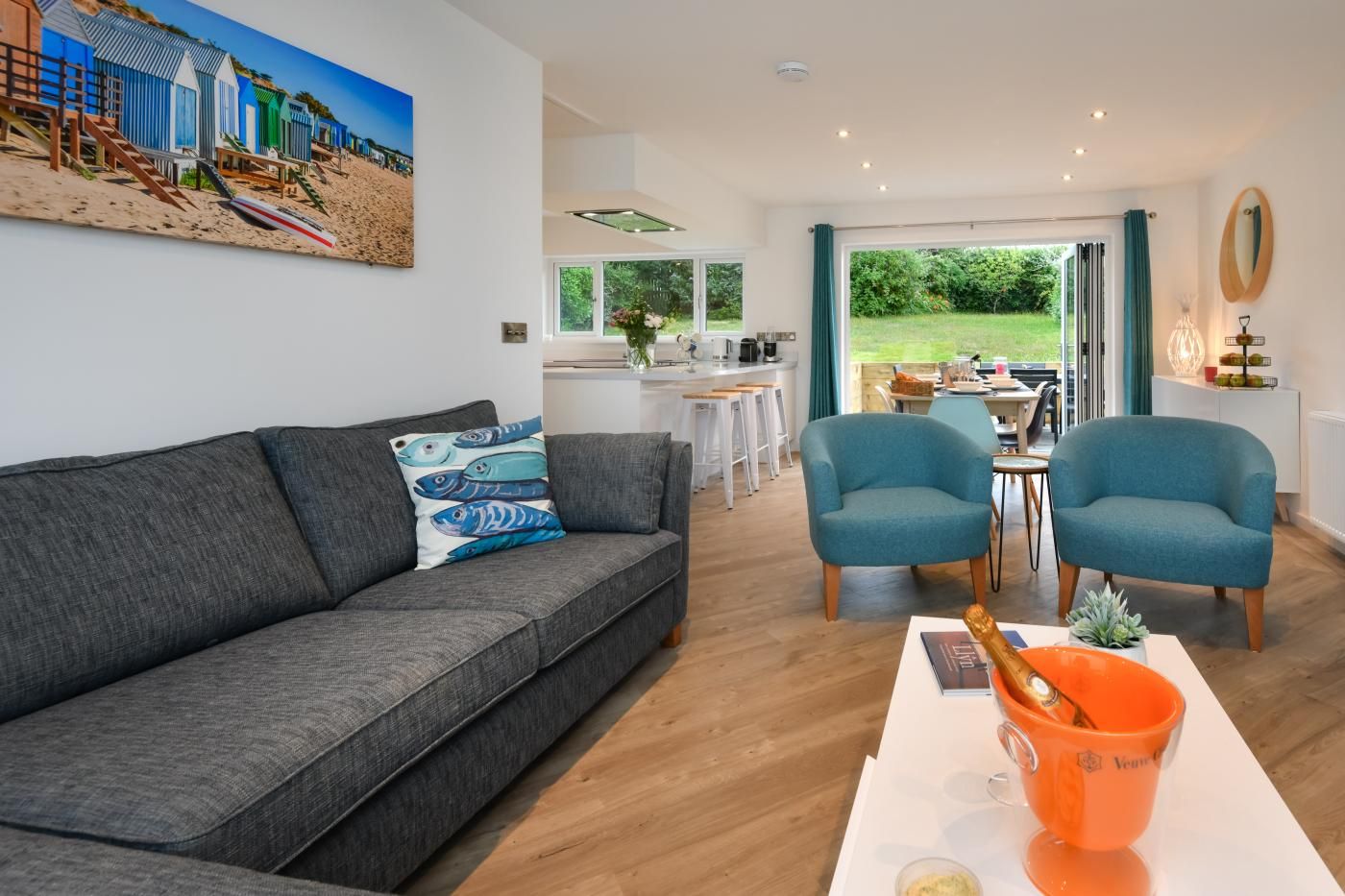 Un salon moderne avec un canapé gris, des fauteuils turquoise, une table basse sur laquelle se trouvent un livre et une boisson, et un tableau représentant une scène de plage.
