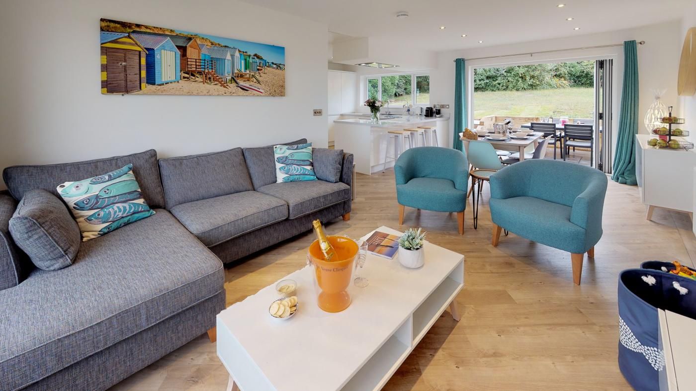 A modern living room with a grey sofa, teal armchairs, a white coffee table with a champagne bottle, and a painting of beach huts.