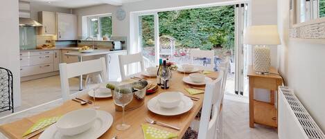 A modern kitchen with white cabinets, a dining area with a wooden table set for four, and a view of a garden through large windows.
