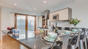 A modern kitchen with a central island, stainless steel appliances, and a dining area with chairs and a table set for two. - 2 Bed in Looe (oc-p00778) (Looe)
