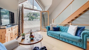 A spacious living room with a large window, a blue sofa, a wooden table with a bowl of fruit, and a television on a wooden cabinet. - 2 Bed in Looe (oc-p00778) (Looe)