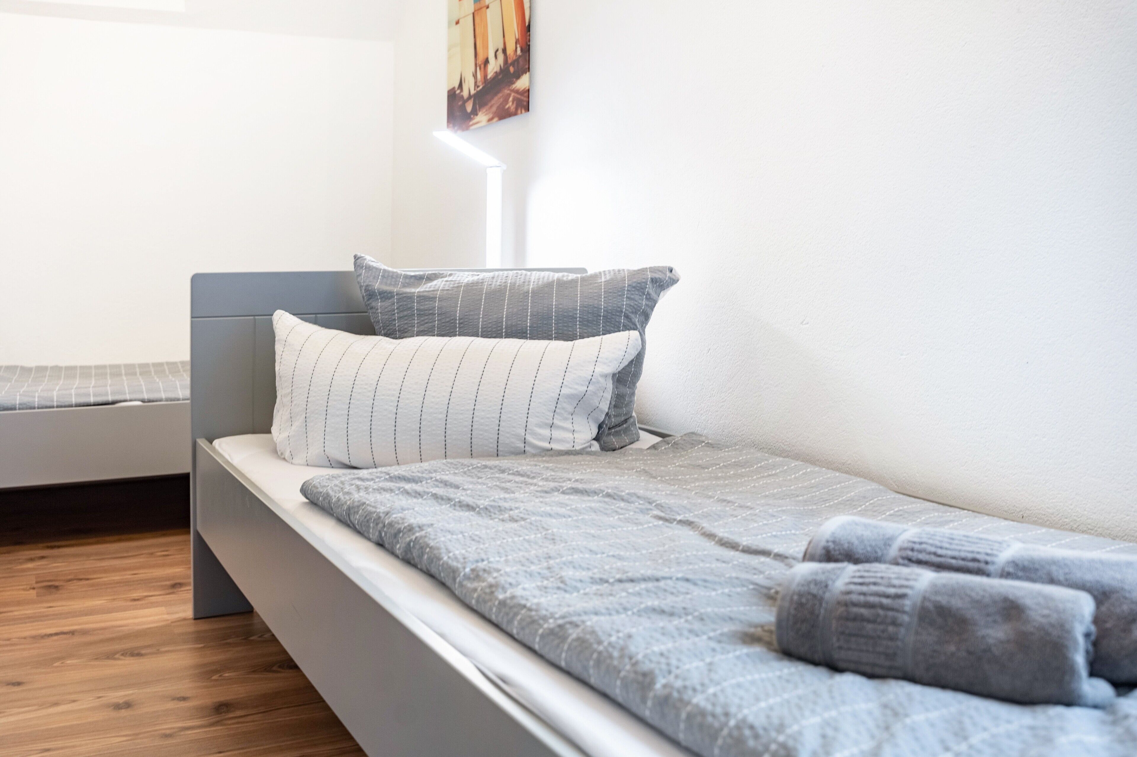Una cama individual con cabecera gris y almohadas, suelo de madera y un cuadro colgado en la pared.