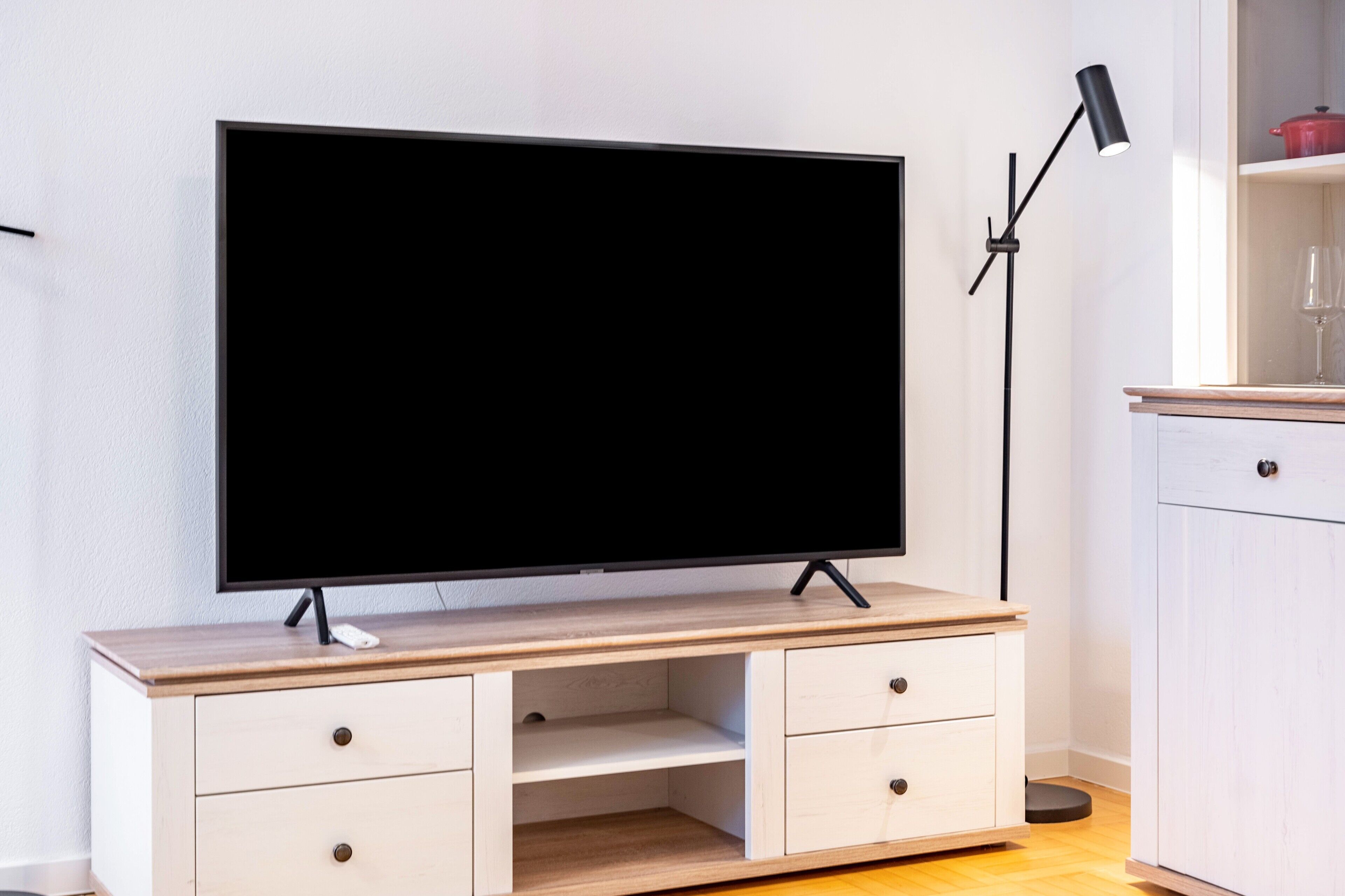 Un televisor pantalla plana sobre un moble de televisió de fusta amb calaixos, una làmpada de peu i un armari blanc amb una olla vermella.